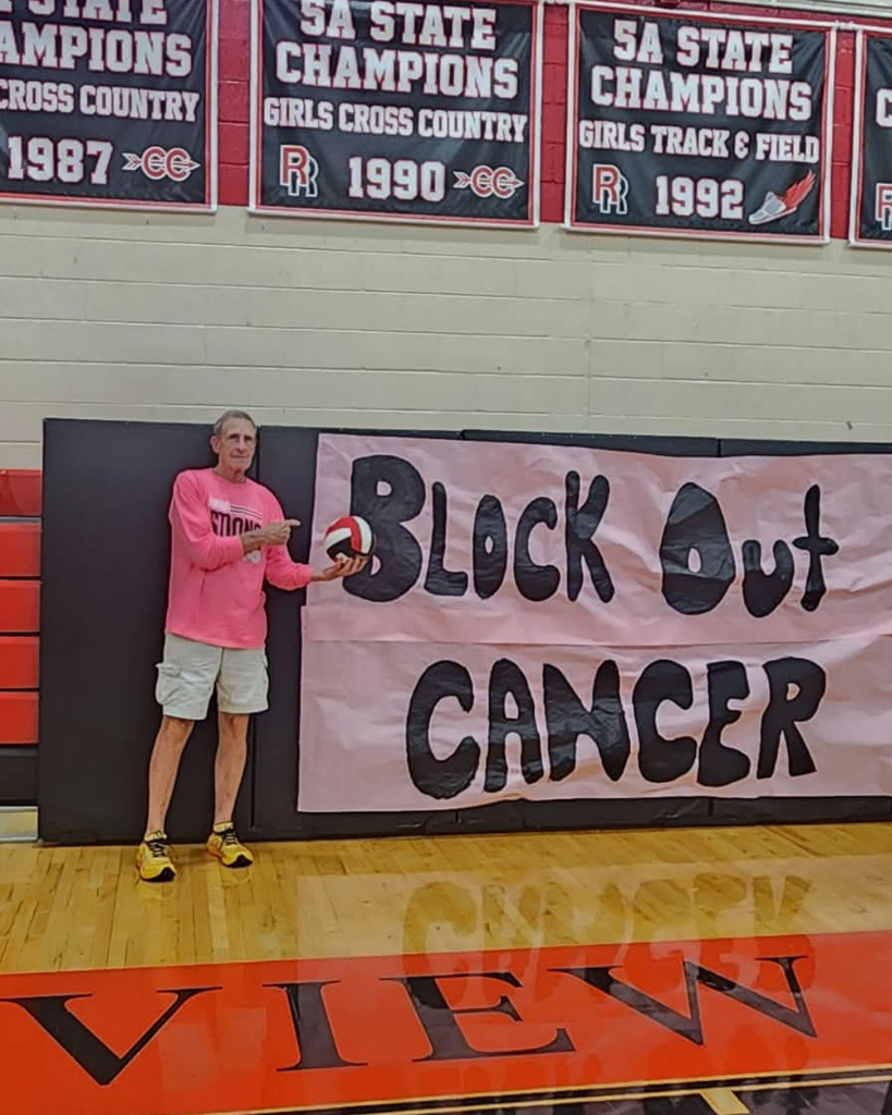 A man in a pink shirt pointing at a volleyball in front of a banner that reads Block Out Cancer in a high school gymnasium.