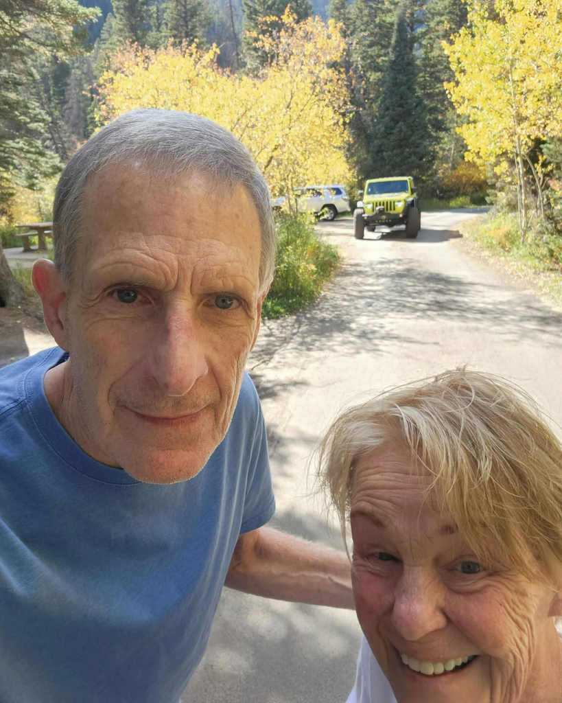 An older man and woman pose for a selfie on a tree-lined road with a lime green Jeep in the background.