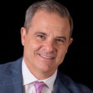 Headshot of a middle-aged man with short hair, wearing a suit jacket, white shirt, and pink-striped tie against a black background, as seen in a podcast about sex after bladder cancer.